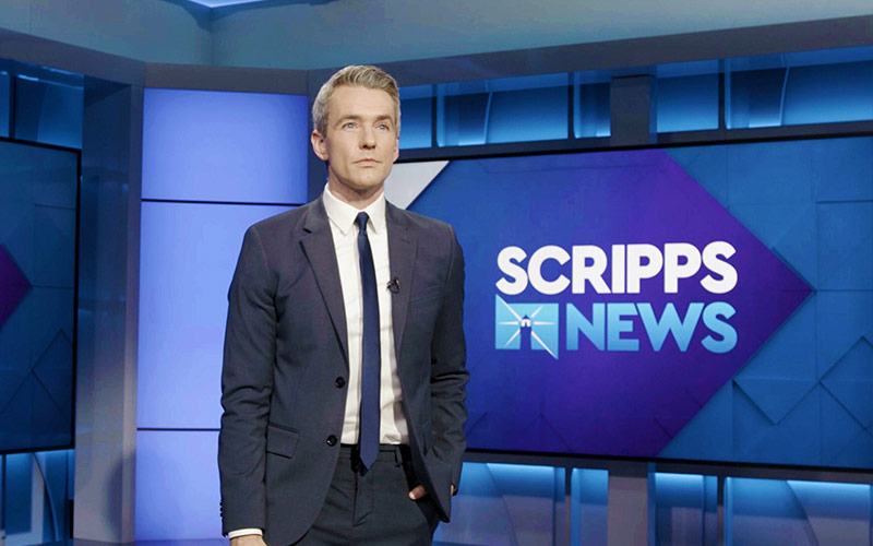 Photo of a man in a navy blue suit standing in front of a screen that says Scripps News